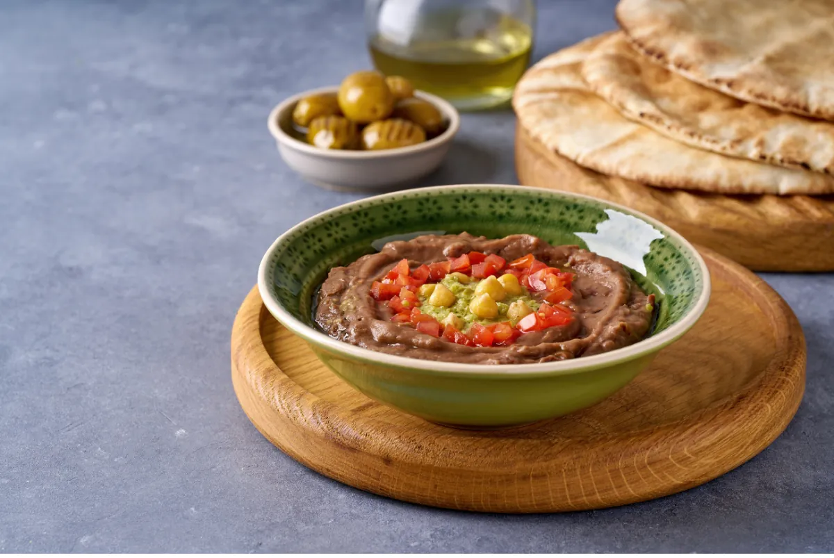 طبق فول مدمس في سلطانية خضراء مزين بالطماطم المقطعة والحمص والأعشاب، وخلفه زيتون وزيت زيتون وخبز.